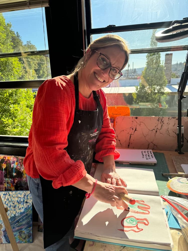 Jen Lyons smiling at her worktable in a sunlit studio, drawing in a sketchbook with an oil pastel