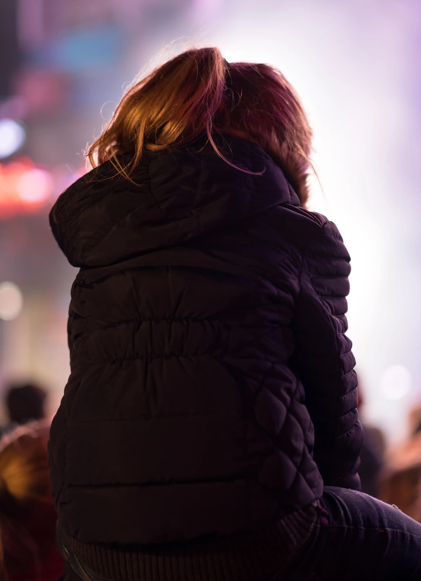 A person in a dark quilted jacket watches a show from behind, lit by soft pink and violet stage lights