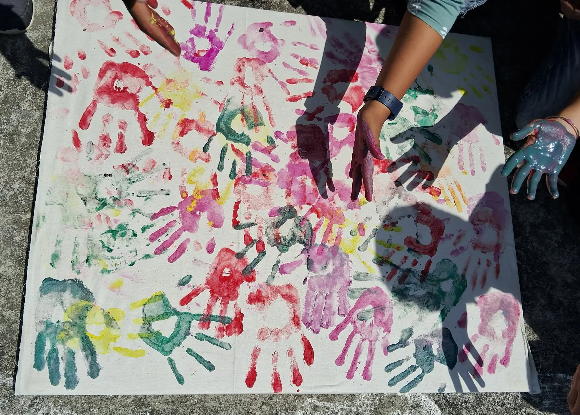 Several paint-covered hands press colourful handprints onto a large white canvas laid on the ground