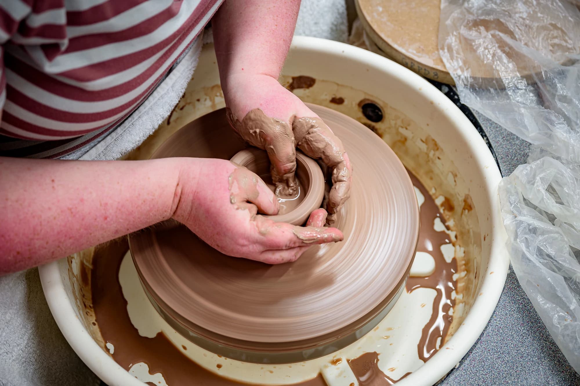 A person in a striped shirt centres a lump of clay on a pottery wheel with wet hands