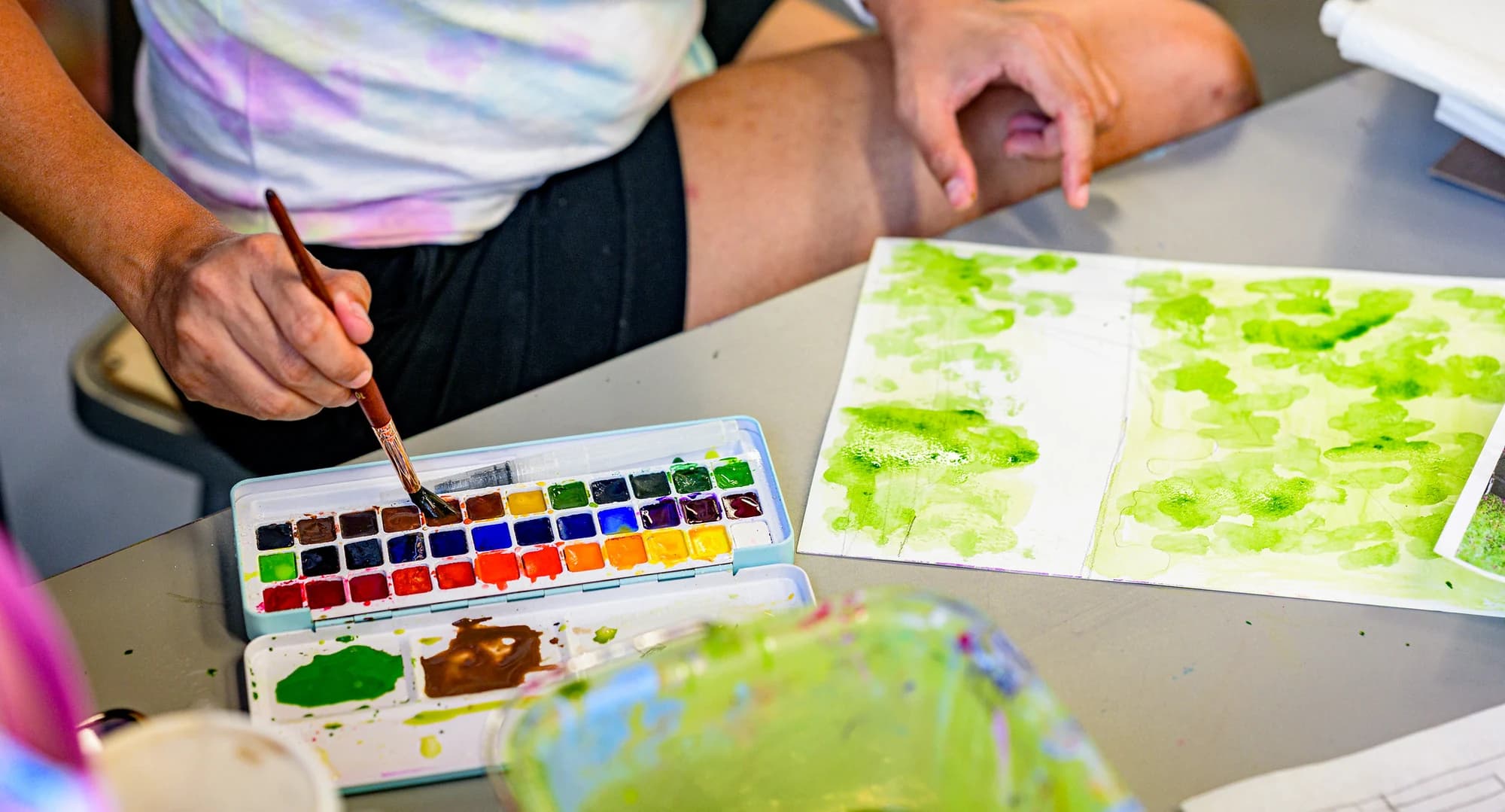 A hand holds a fine brush over a watercolour pan next to sheets of fresh green paint swatches