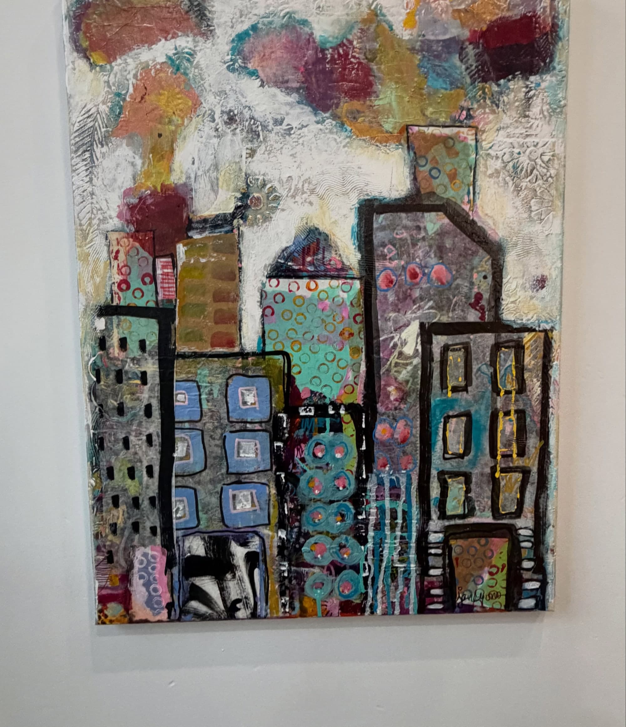 A colourful painting of city buildings in layered blues, oranges and greens, hanging on the gallery wall