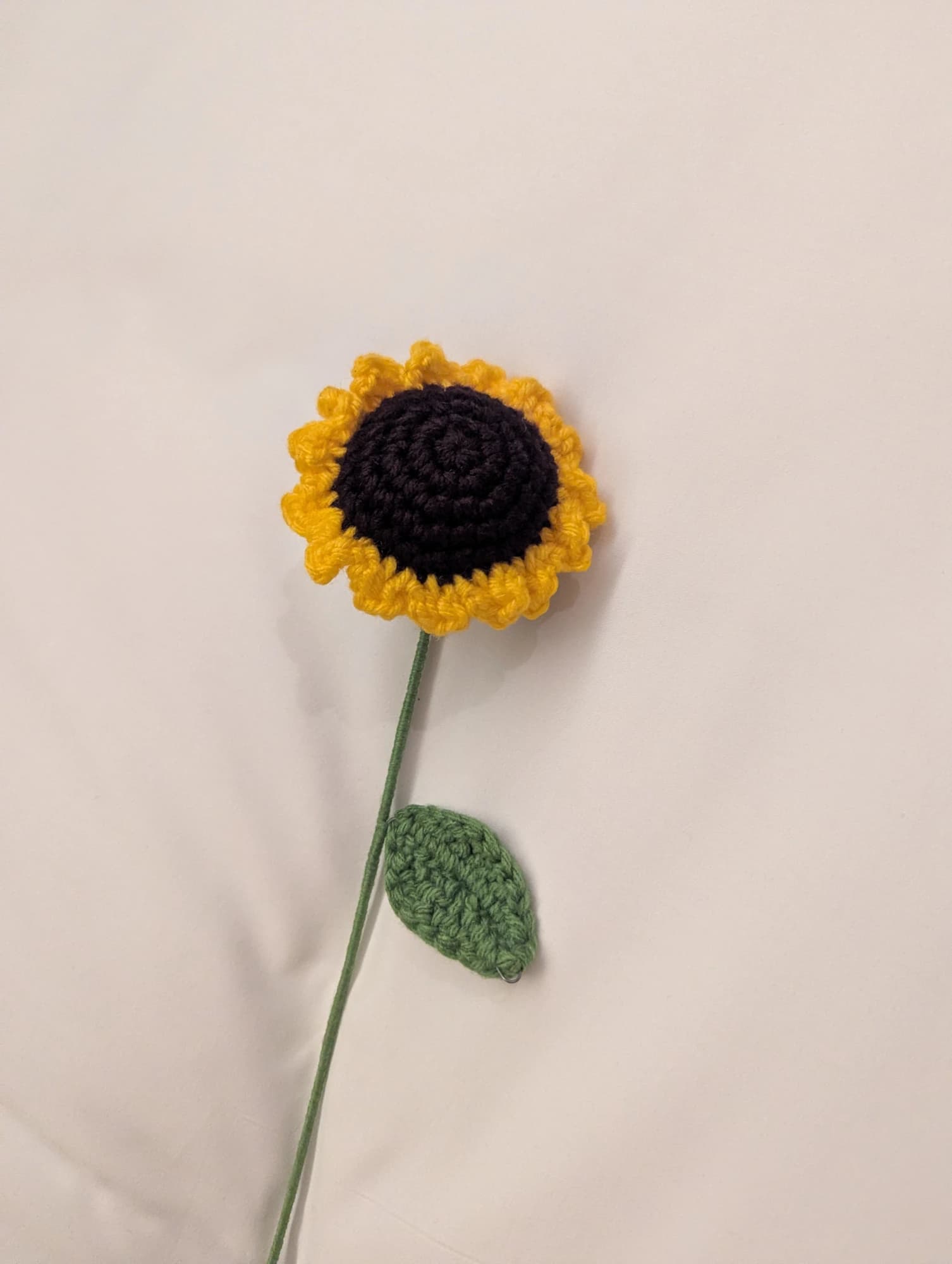 A single crocheted sunflower with a dark centre, yellow petals and a green leaf lies on a soft cream fabric