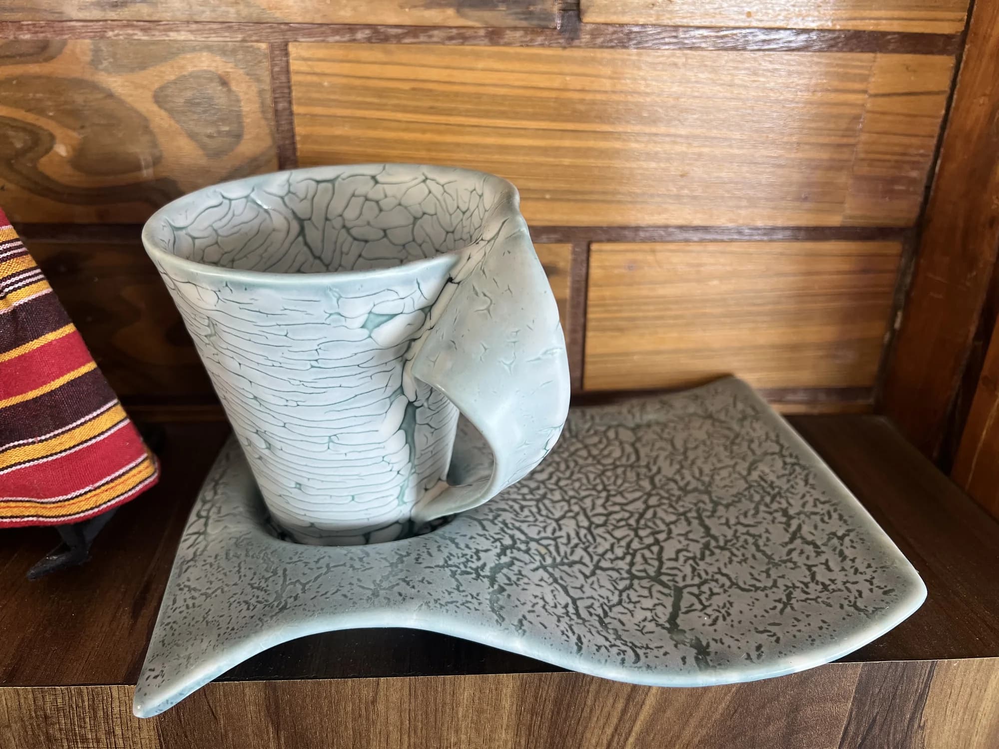 A pale blue ceramic cup with a crackled glaze rests on a wavy matching tray on a wooden shelf