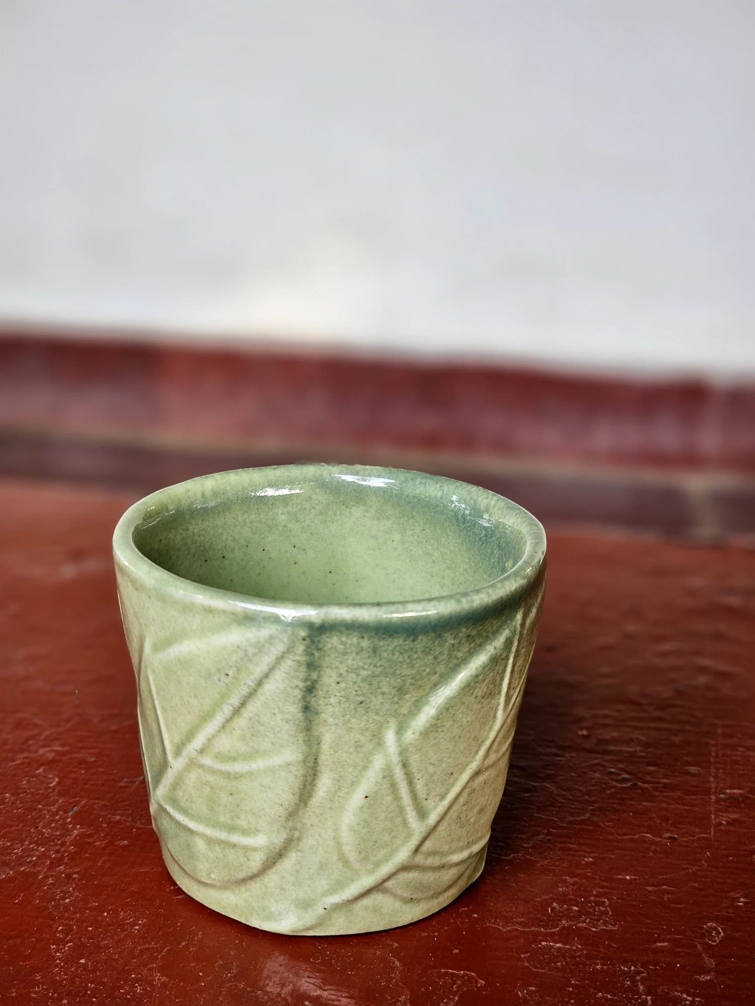 A small sage green ceramic cup with a carved leaf pattern stands on a dark red surface against a pale wall