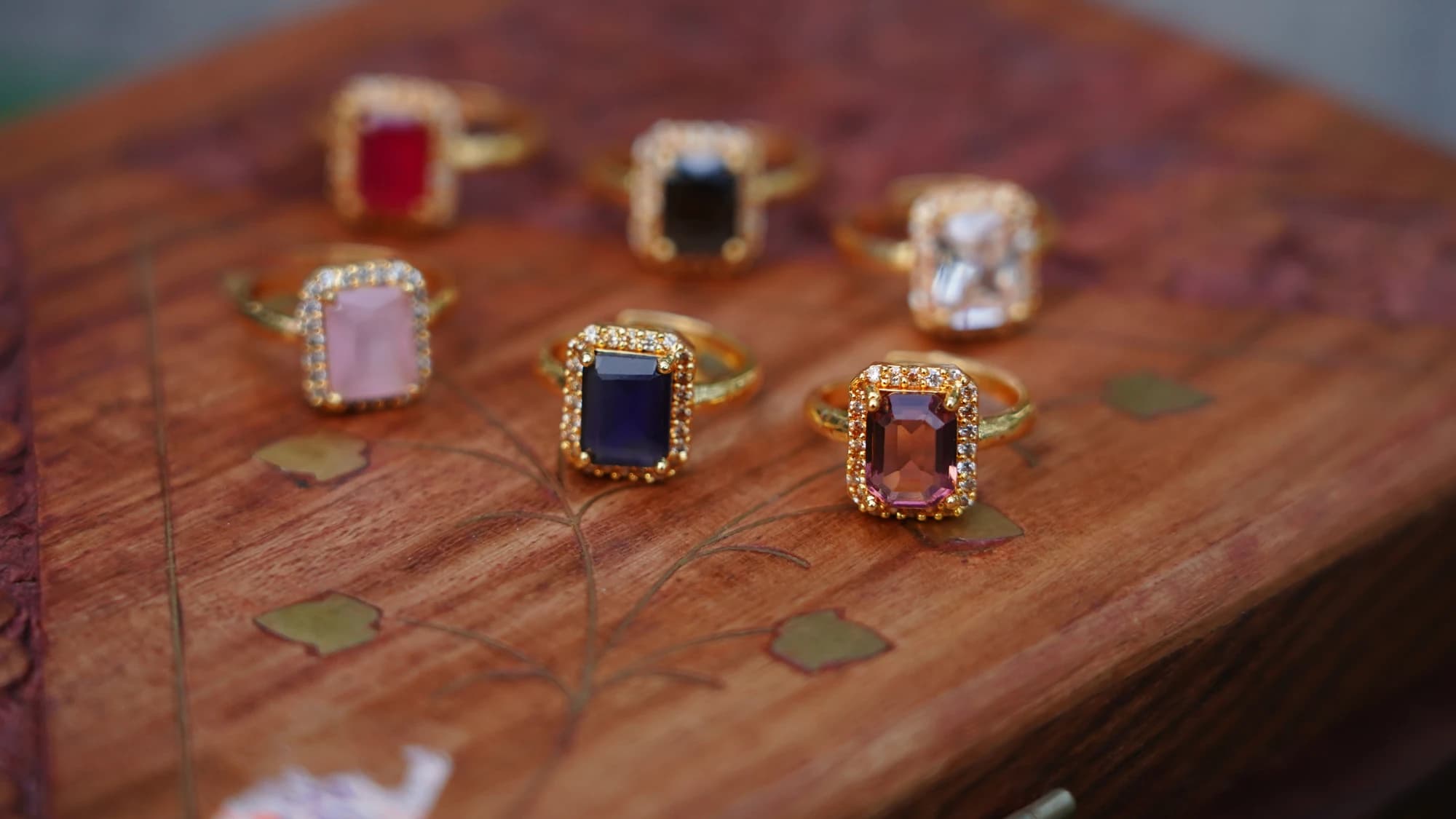 Six gold rings set with pink, red, blue and clear stones lie scattered on a polished wooden board