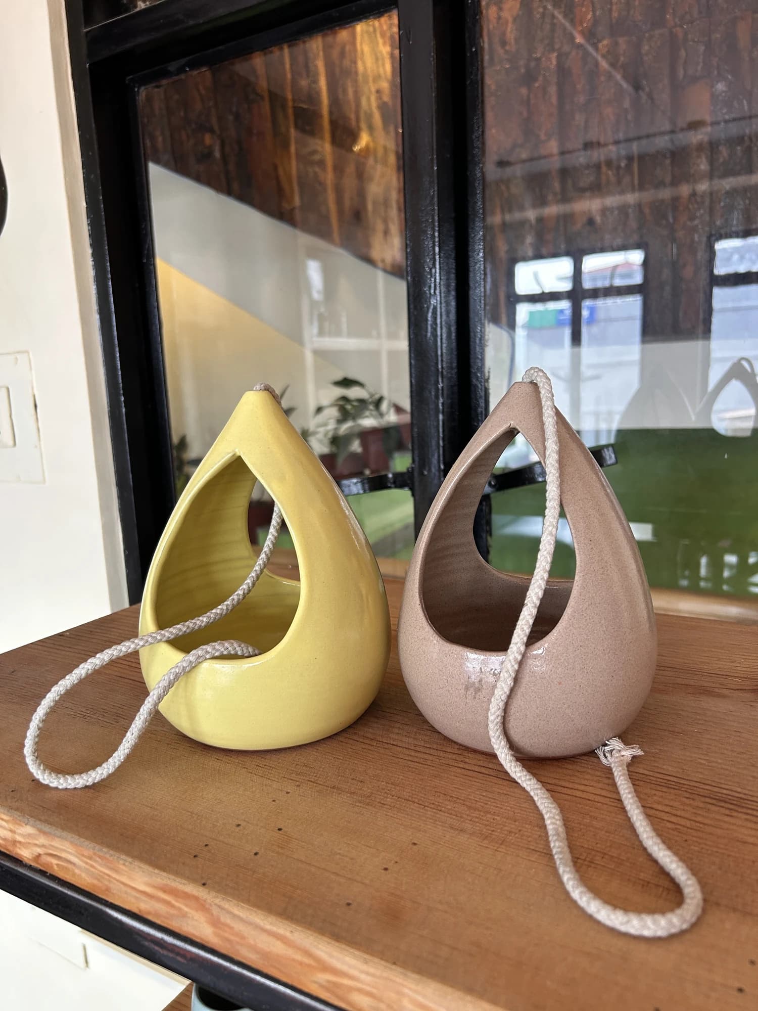 Two teardrop shaped ceramic hanging planters, one yellow and one blush pink, stand on a wooden shelf with their cords coiled