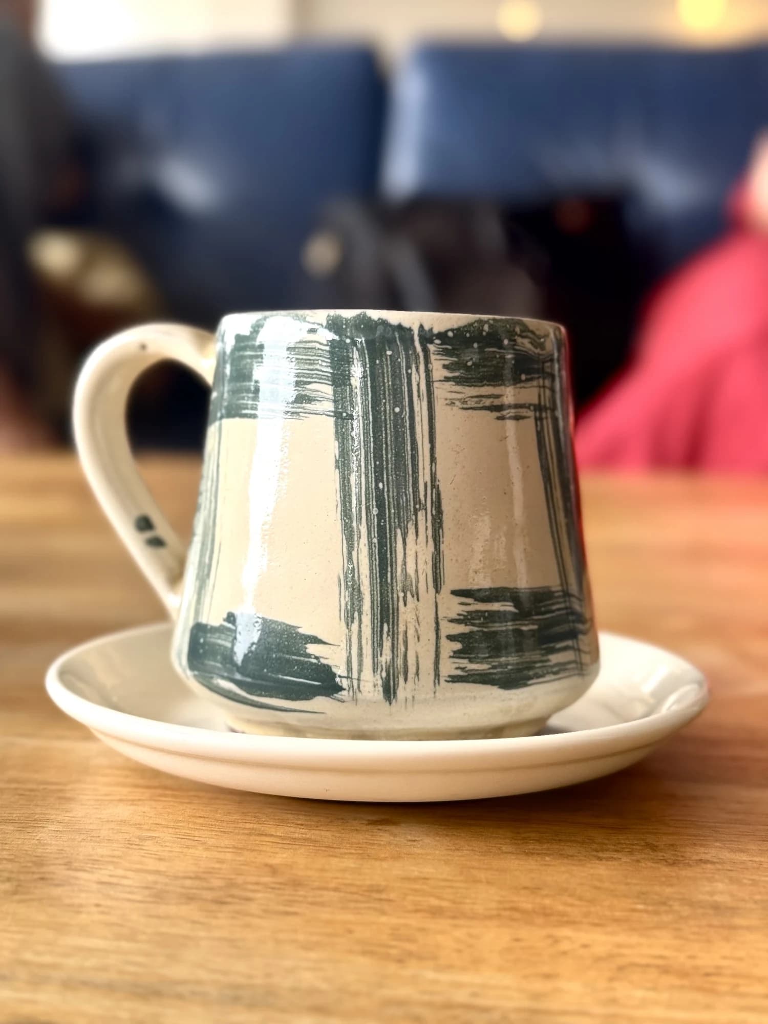 A handmade cream mug with loose grey brushstroke glaze sits on a matching saucer on a wooden table